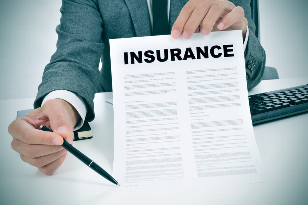 A person in a suit holds out a document titled INSURANCE and points to it with a pen, suggesting a discussion about an insurance policy.