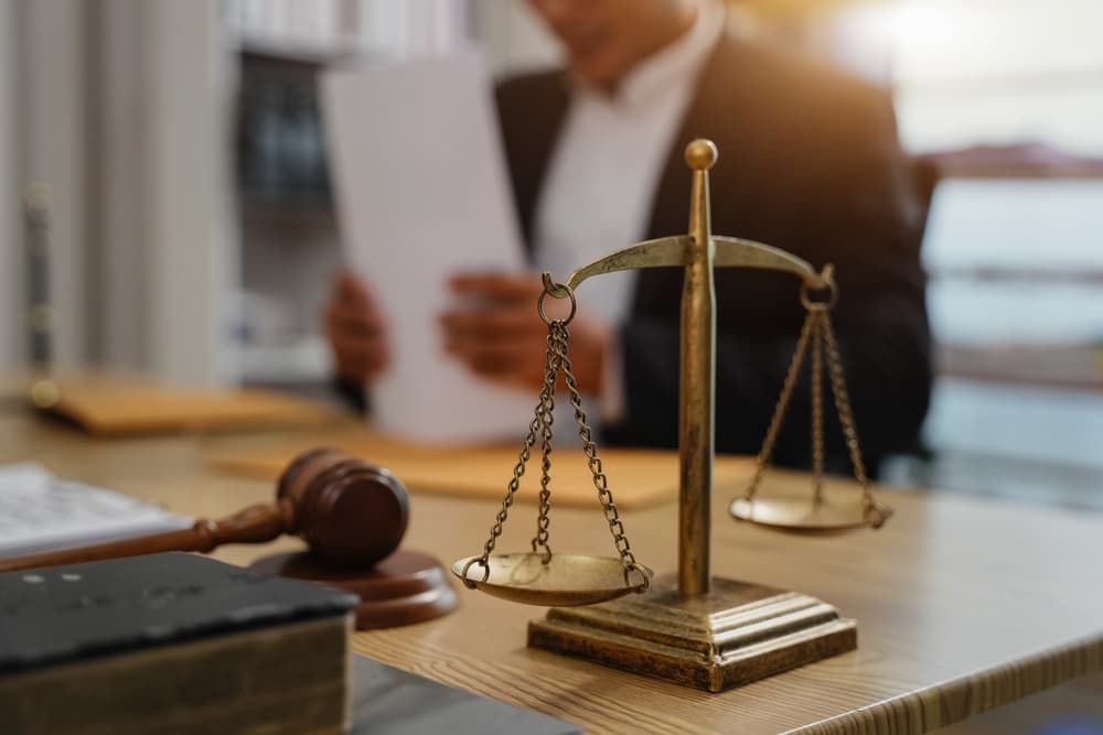 Close-up of a brass justice scale and judge’s gavel on a desk with a blurred lawyer reading documents in the background.