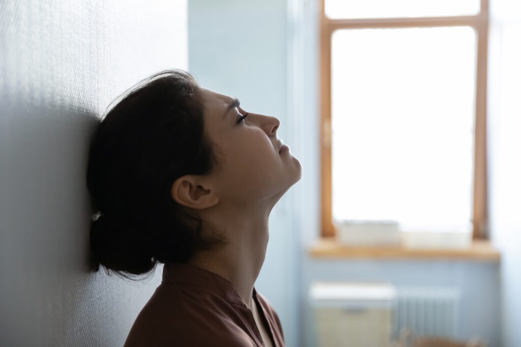 Woman leaning against a wall with eyes closed and head tilted back, appearing emotionally overwhelmed and showing signs of post-traumatic stress disorder (PTSD).