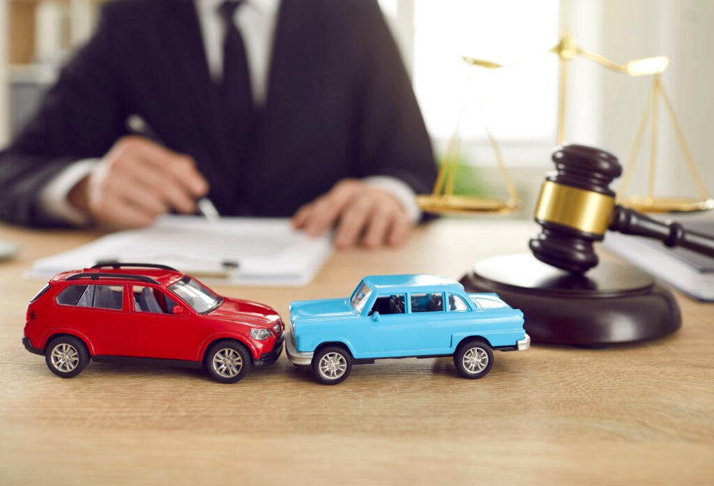 San Jose Car Accident  Lawyer. A red car and a blue car on a desk with a gavel and scales. Lawyer writing in the background.